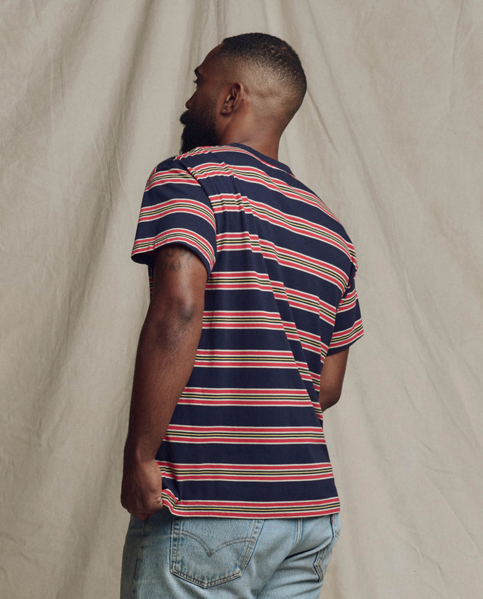 A man stands facing away, wearing THE GREAT. SP26 SEASONAL CORE The Men's Boxy Crew in Scout Stripe and light blue jeans, set against a beige fabric backdrop.
