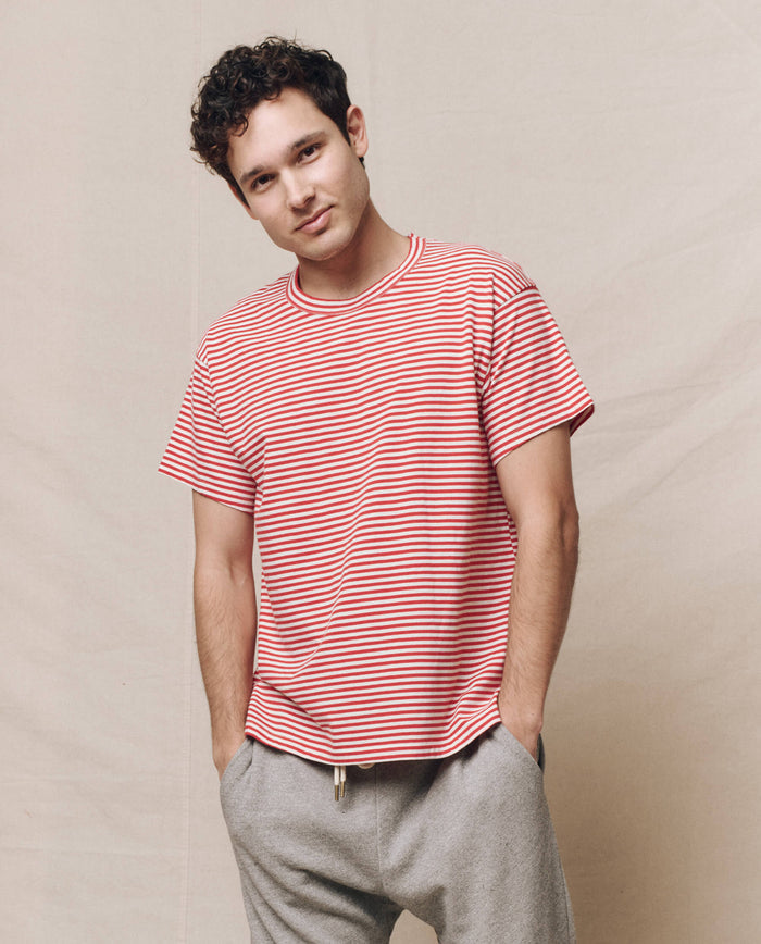 A person wearing THE GREAT. SP25 POLO LOUNGE MEN's The Men's Boxy Crew in Sun Cherry Stripe and gray pants stands against a beige background.