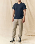 A person in THE GREAT. FALL 23 MEN The Men's Boxy Crew -- True Navy stands before a beige fabric backdrop, paired with gray jogger sweatpants and black high-top sneakers.