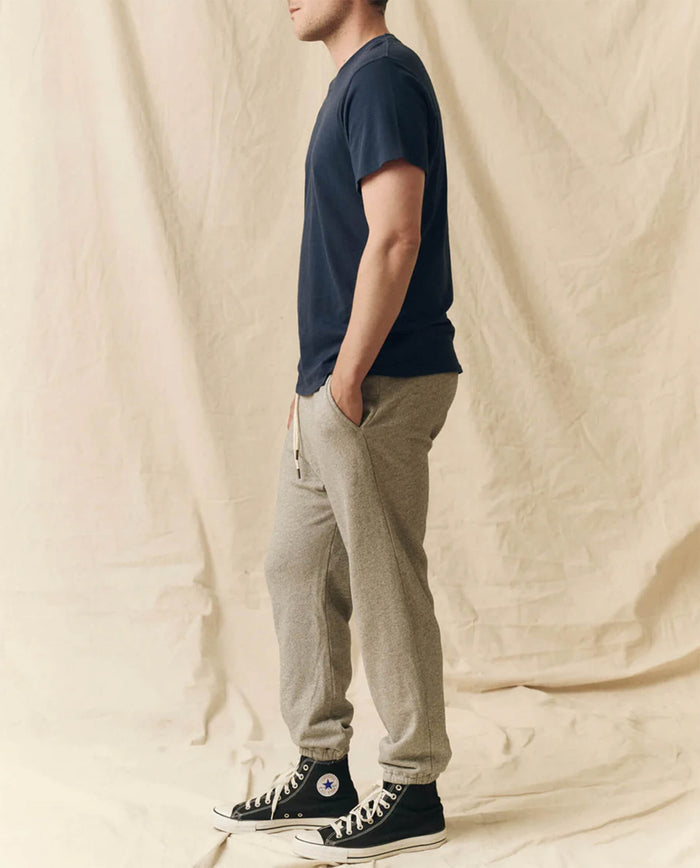 A person stands in profile, hands in pockets, wearing THE GREAT. FALL 23 MEN The Men's Boxy Crew. -- True Navy, gray sweatpants, and black high-top sneakers against a beige fabric backdrop.