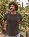 A man with curly hair and a beard is standing outdoors in front of plants, wearing The Men's Boxy Crew in Washed Black by THE GREAT. MAN and jeans, with his hands behind his back.