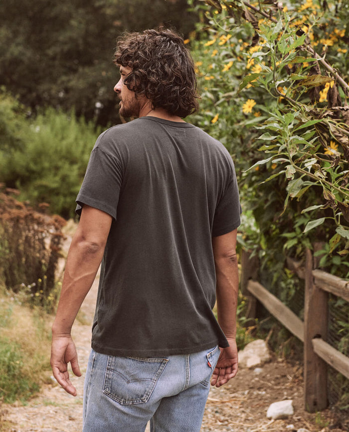 A person with curly hair and a beard stands on a dirt path next to a wooden fence and greenery, facing away from the camera. They are wearing The Men's Boxy Crew in washed black by THE GREAT. MAN, which is a timeless wardrobe staple, paired with light blue jeans.