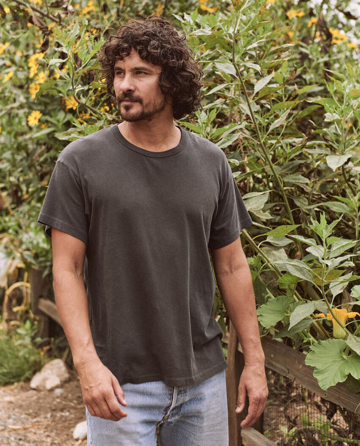 A man with curly hair and a beard stands outdoors, wearing The Men's Boxy Crew in Washed Black by THE GREAT. MAN and light jeans, with green leafy plants and yellow flowers in the background.