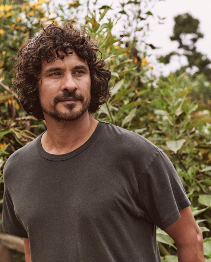 A man with curly hair and a beard stands outside, wearing The Men's Boxy Crew in washed black by THE GREAT. MAN. He is looking off into the distance with greenery in the background.