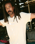 A man with long dreadlocks and a beard, wearing The Men's Boxy Crew in washed white by THE GREAT. MAN, stands with his arms slightly raised in an outdoor setting.