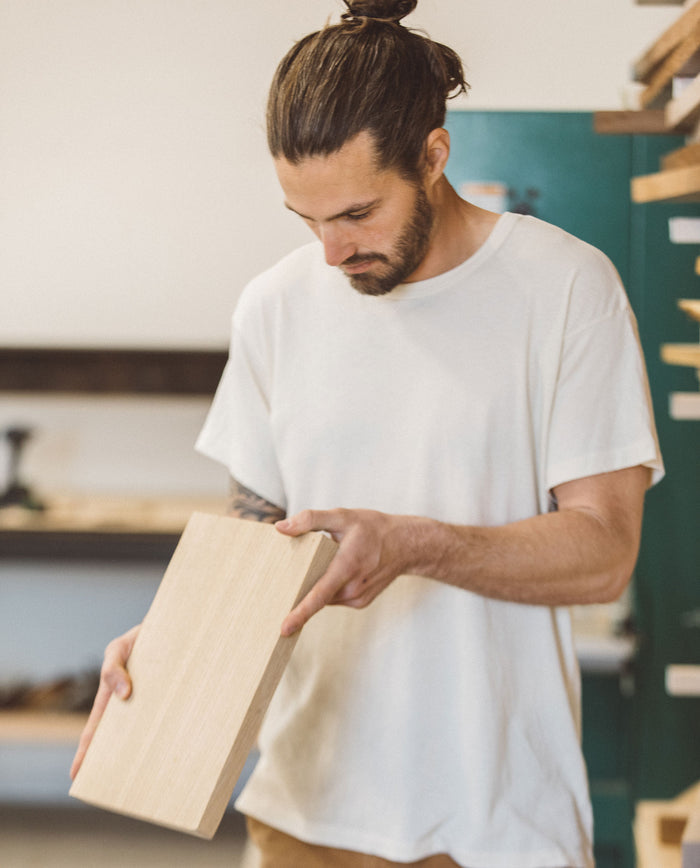 A person in The Men's Boxy Crew in Washed White by THE GREAT. MAN inspects a wooden plank in a workshop environment.