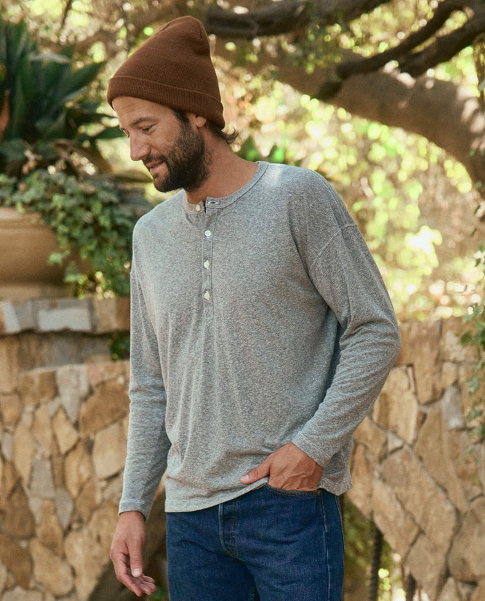 A man wearing The Men's Boxy Henley in Heather Grey by THE GREAT. HOL 22 MEN, blue jeans, and a brown beanie stands outdoors near a stone wall and trees in Los Angeles.