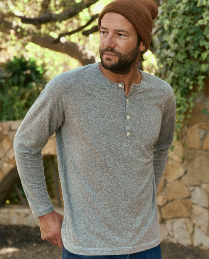 Man wearing THE GREAT. HOL 22 MEN's The Men's Boxy Henley in Heather Grey and a brown beanie, standing outdoors in front of a stone wall and foliage in Los Angeles.