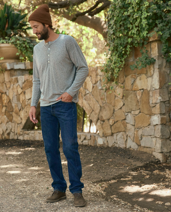 A man in a heather grey Men's Boxy Henley from THE GREAT. HOL 22 MEN, blue jeans, brown beanie, and shoes stands leisurely on a dirt path beside a stone wall with greenery in Los Angeles.