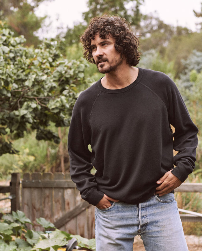 A man with curly hair and a beard, wearing The Men's College Sweatshirt by THE GREAT. MAN in Almost Black and jeans, stands outdoors near a wooden fence and greenery.