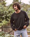 A man with curly hair and a beard, wearing The Men's College Sweatshirt by THE GREAT. MAN in Almost Black and jeans, stands outdoors near a wooden fence and greenery.