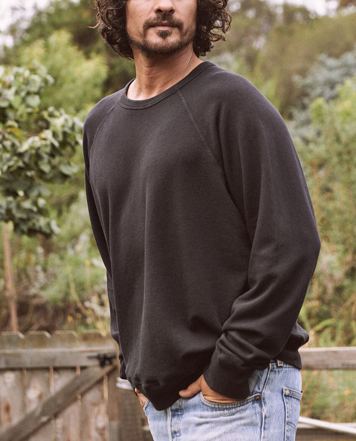 A man with curly hair and a beard is standing outdoors, wearing The Men's College Sweatshirt in Almost Black by THE GREAT. MAN and jeans. Trees and a wooden fence are visible in the background.