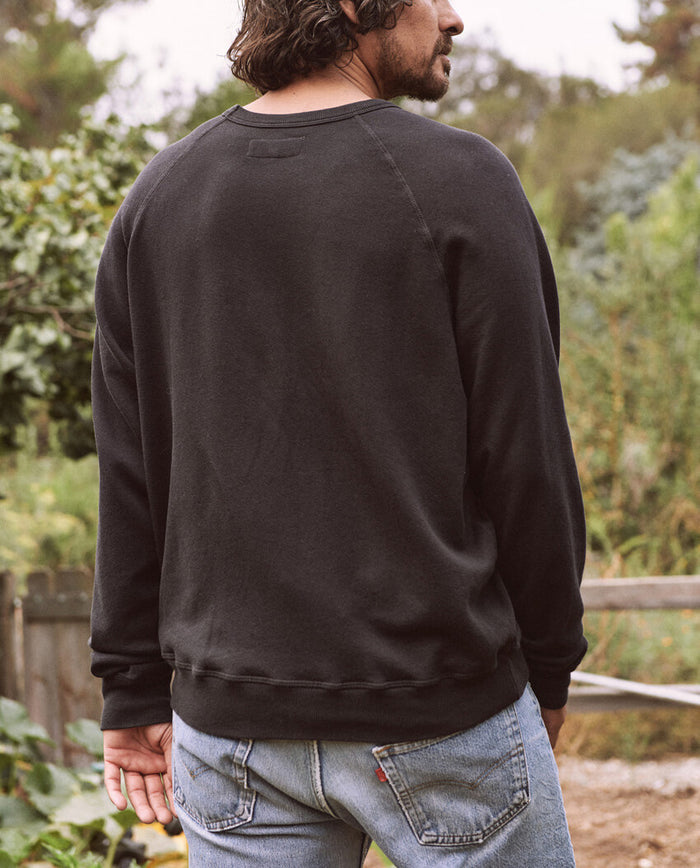 A man with wavy hair stands outdoors wearing The Men's College Sweatshirt in Almost Black from THE GREAT. MAN, paired with jeans. His back is facing the camera, and there is greenery in the background.