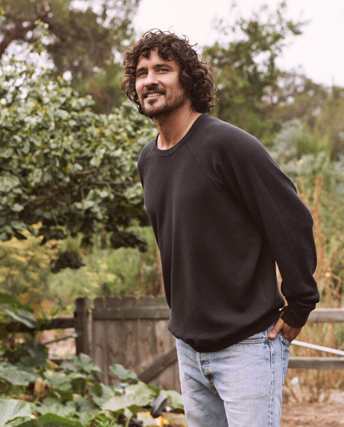A man with curly hair and a beard is standing outdoors near greenery and a wooden fence, smiling and looking up. He's wearing The Men's College Sweatshirt in Almost Black by THE GREAT. MAN, paired with jeans.