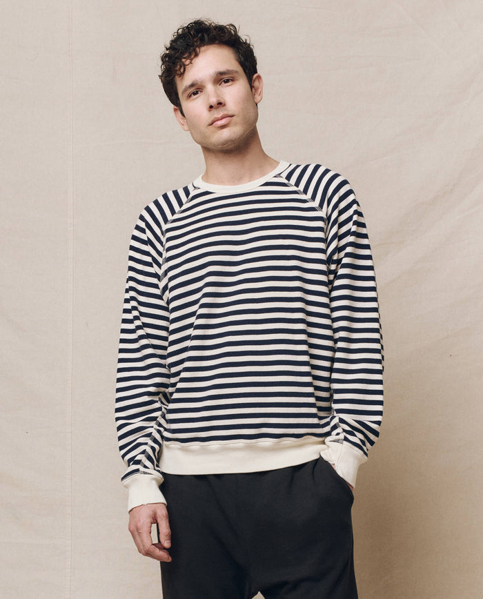 Wearing THE GREAT. SP25 POLO LOUNGE MEN's The Men's College Sweatshirt in Navy Sailor Stripe, paired with dark pants, a person stands against a plain background.