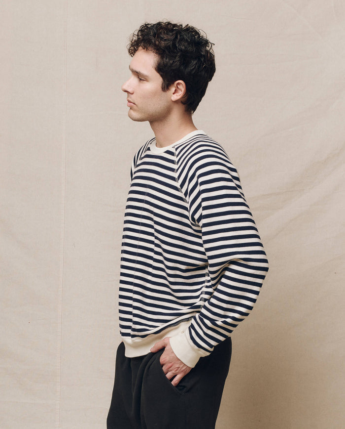 A person with curly hair, wearing The Men's College Sweatshirt in Navy Sailor Stripe by THE GREAT. SP25 POLO LOUNGE MEN, stands sideways against a plain backdrop with hands in pockets, effortlessly embodying stylish collegiate men's clothing.