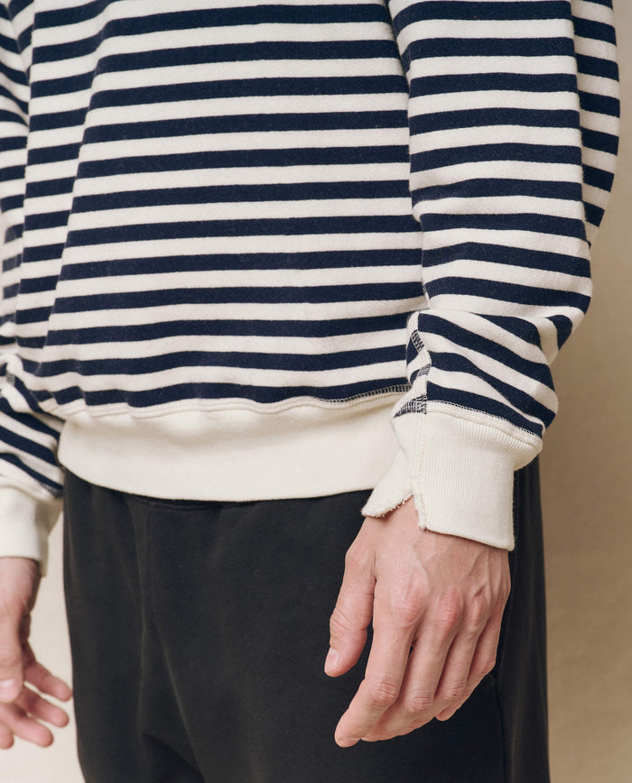A person wearing THE GREAT. SP25 POLO LOUNGE MEN's The Men's College Sweatshirt in Navy Sailor Stripe with dark pants stands against a neutral background, their hands visible at their sides.