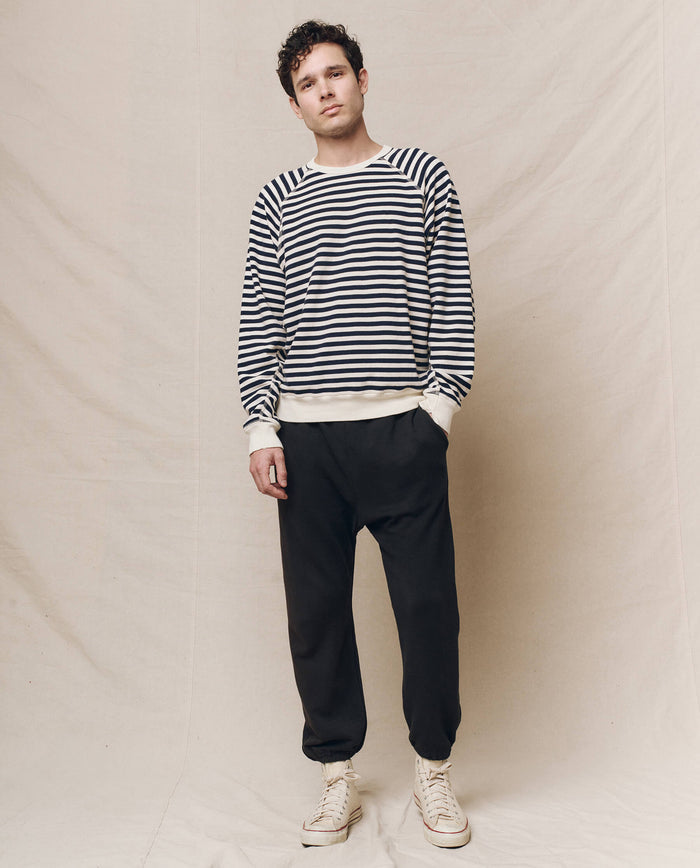 A person stands on a fabric-covered floor, wearing THE GREAT. SP25 POLO LOUNGE MEN's ensemble: The Men's College Sweatshirt in Navy Sailor Stripe, black pants, and light-colored sneakers. Their neutral expression complements the casual college vibe with hands resting by their sides.