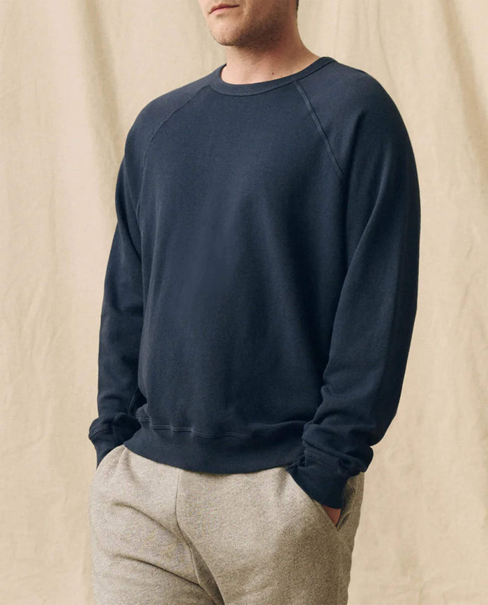 A person in The Men's College Sweatshirt -- True Navy by THE GREAT. FALL 23 MEN and light pants stands with hands in pockets against a beige fabric background.