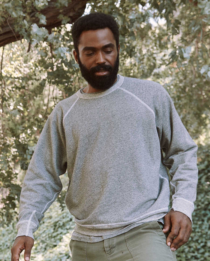 A bearded man stands outdoors amid lush greenery, clad in the varsity grey Men's College Sweatshirt by THE GREAT. MAN.