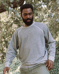 A bearded man stands outdoors amid lush greenery, clad in the varsity grey Men's College Sweatshirt by THE GREAT. MAN.