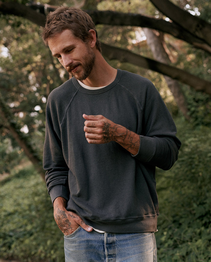 A man wearing The Men's College Sweatshirt in washed black by THE GREAT. MAN and jeans stands outdoors, looking down with one hand in his pocket and the other gesturing slightly. He has tattoos on his hands and arms. Trees can be seen in the background. The sweatshirt is made in the U.S.A., ensuring quality craftsmanship.