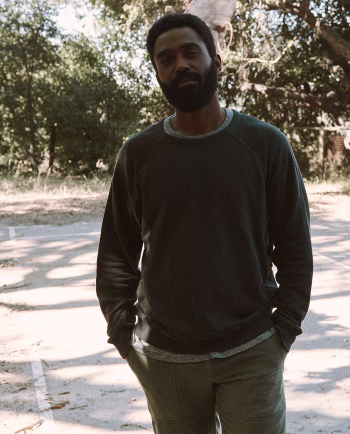 A man stands outdoors on a sunlit, tree-lined path, wearing THE GREAT. MAN's washed black, vintage Men's College Sweatshirt and light-colored pants. Hands in his pockets, he looks directly at the camera.