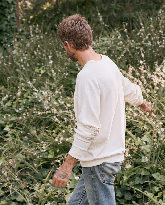 A person with short hair wearing The Men's College Sweatshirt in washed white by THE GREAT. MAN and blue jeans walks through a lush, green garden.