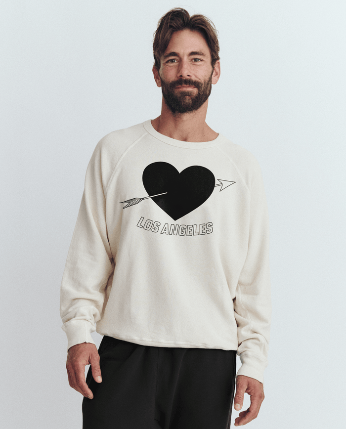 A man wears The Men's College Sweatshirt by THE GREAT. SP25 I LOVE LA, showcasing a washed white design with a Los Angeles heart graphic and arrow, smiling against a plain background.
