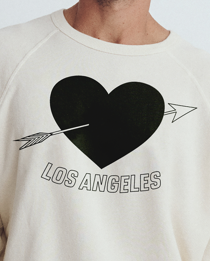 Someone wears The Men's College Sweatshirt by THE GREAT. SP25 I LOVE LA, a washed white design with an LA heart graphic featuring a black heart pierced by an arrow and "Los Angeles" text.
