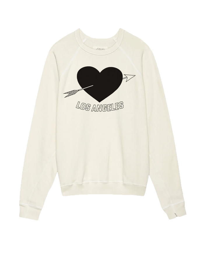 The Men's College Sweatshirt by THE GREAT. SP25 I LOVE LA features a washed white design with an LA heart graphic, depicting a black heart pierced by an arrow and the text "Los Angeles" across it.