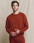 A man with curly hair models THE GREAT. F25 MENS The Men's College Sweatshirt in Solid -- Brick, along with matching pants, standing before a beige fabric backdrop.