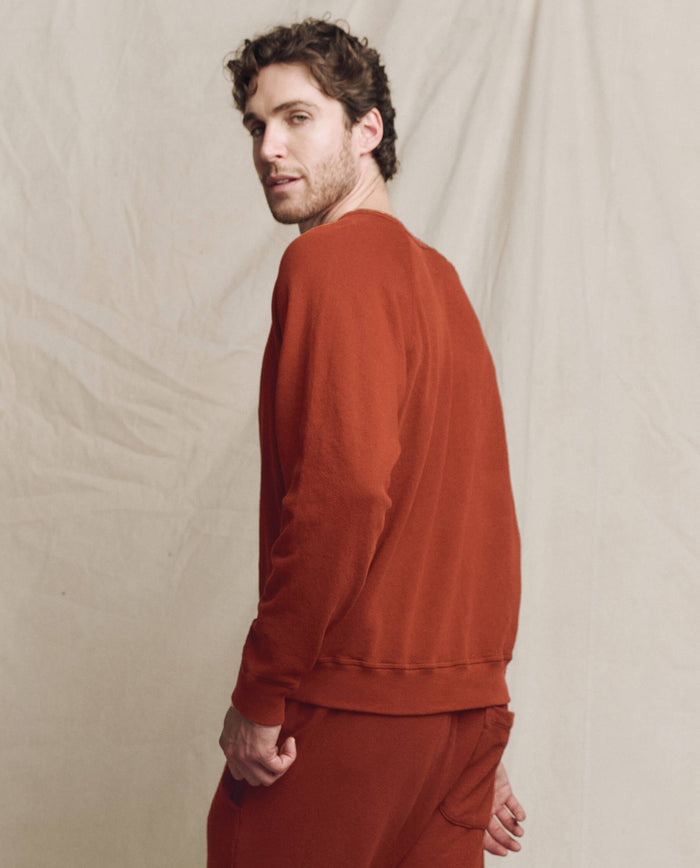 A man with curly hair wears THE GREAT. F25 MENS The Men's College Sweatshirt, Solid -- Brick, and matching pants, looking over his shoulder against a beige fabric backdrop.