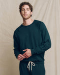 A man wears THE GREAT. F25 MENS The Men's College Sweatshirt in Solid Dark Slate with matching pants featuring white drawstrings, standing against a beige fabric backdrop.