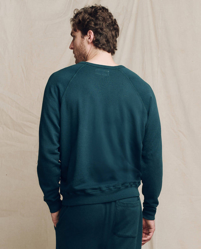 A man with curly hair, seen from behind, wears THE GREAT. F25 MENS The Men's College Sweatshirt in solid Dark Slate with matching pants, standing against a light beige background.