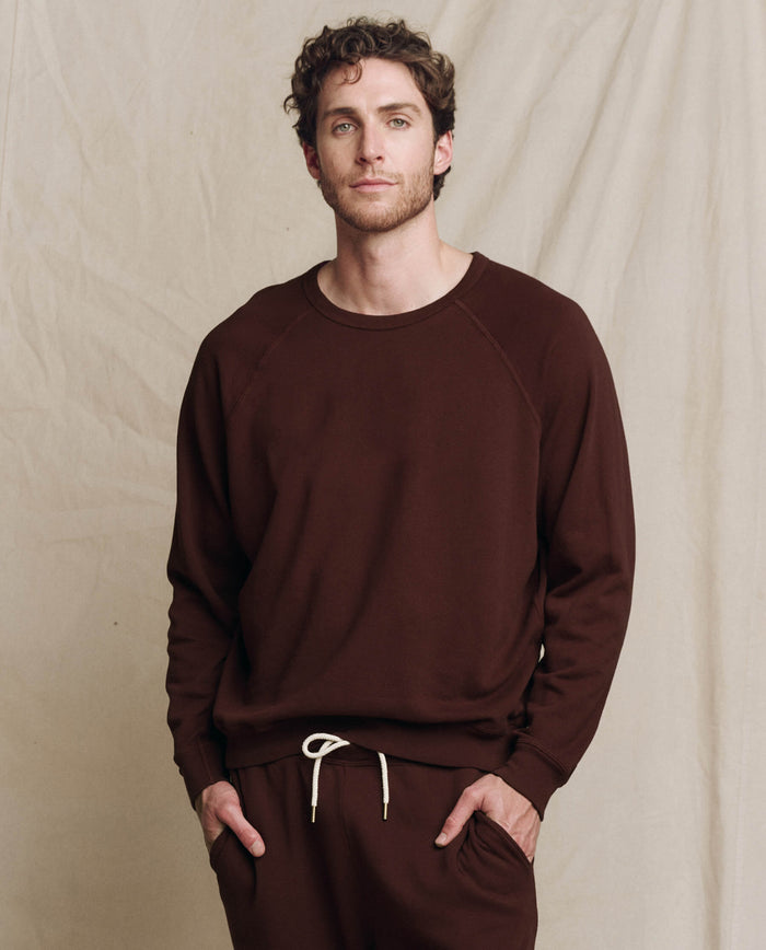 A man wearing THE GREAT. F25 MENS The Men's College Sweatshirt in Driftwood stands before a beige fabric backdrop, looking at the camera with a neutral expression.