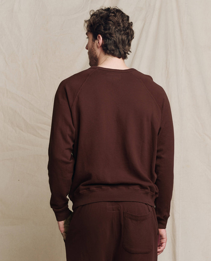 A man with curly hair, wearing THE GREAT. F25 MENS The Men's College Sweatshirt in Solid Driftwood, stands facing away from the camera against a light-colored fabric backdrop.