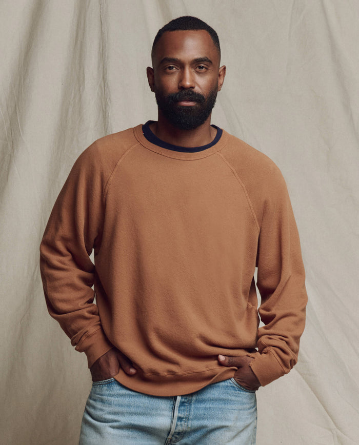A man with a short beard and hair stands against a beige backdrop, wearing THE GREAT. SP26 SEASONAL CORE "The Men's College Sweatshirt" in Rust and light blue jeans, his hands tucked in his pockets.