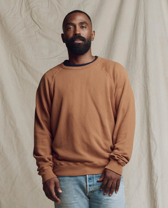 A man with a beard stands in front of a beige fabric backdrop, wearing THE GREAT. SP26 SEASONAL CORE The Men's College Sweatshirt in rust and light blue jeans.