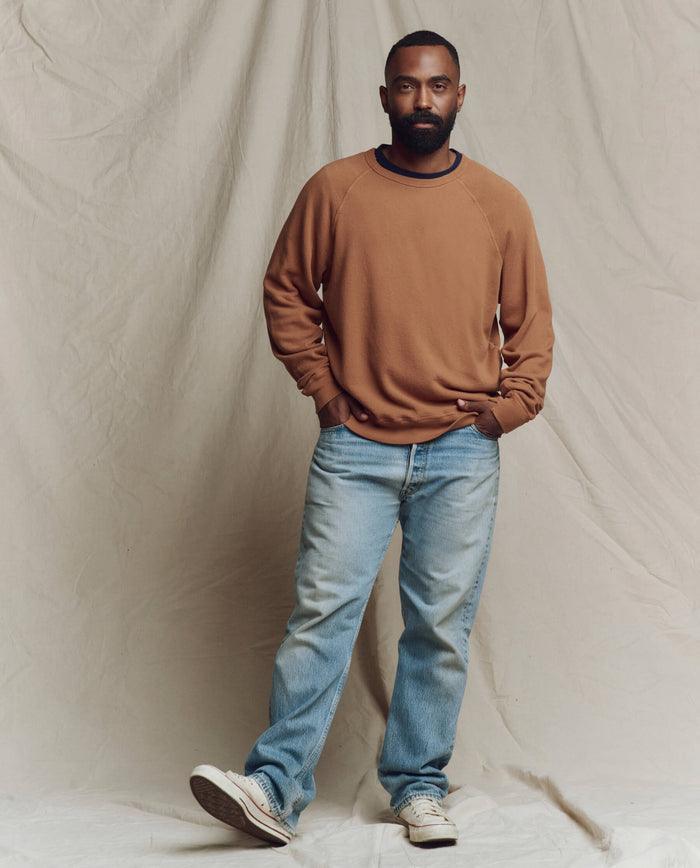 A man stands before a beige fabric backdrop, wearing THE GREAT. SP26 SEASONAL CORE The Men's College Sweatshirt in Rust, paired with light blue jeans and white sneakers, his hands tucked in his pockets.