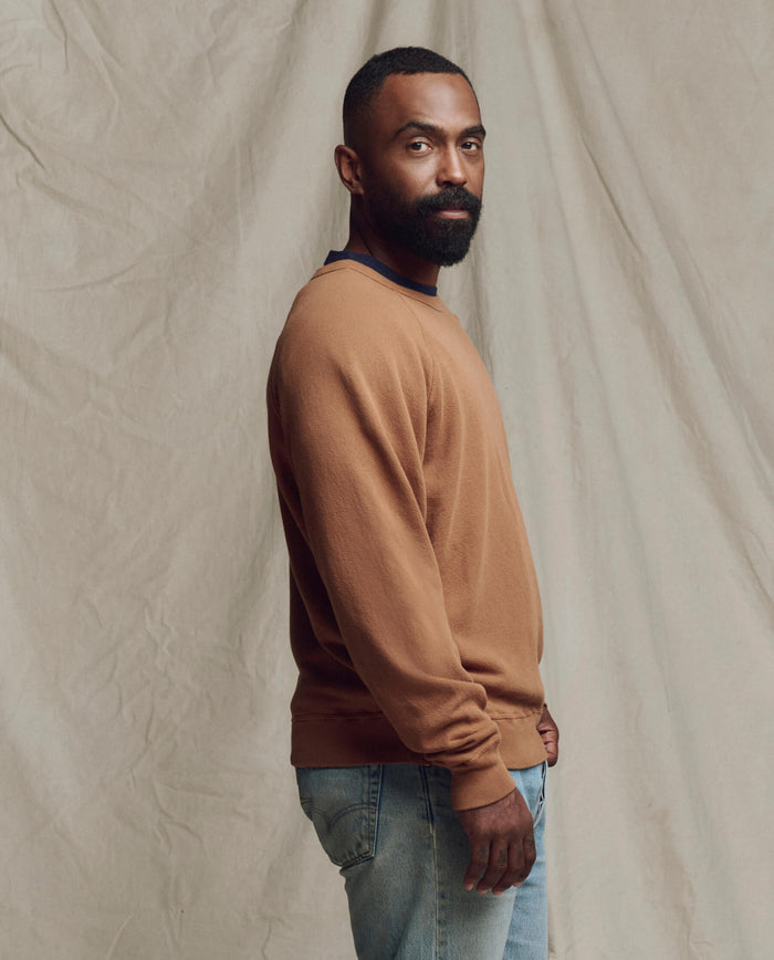 A bearded man stands sideways before a neutral fabric backdrop, wearing THE GREAT.'s Men's College Sweatshirt in Rust from the SP26 SEASONAL CORE and light blue jeans.