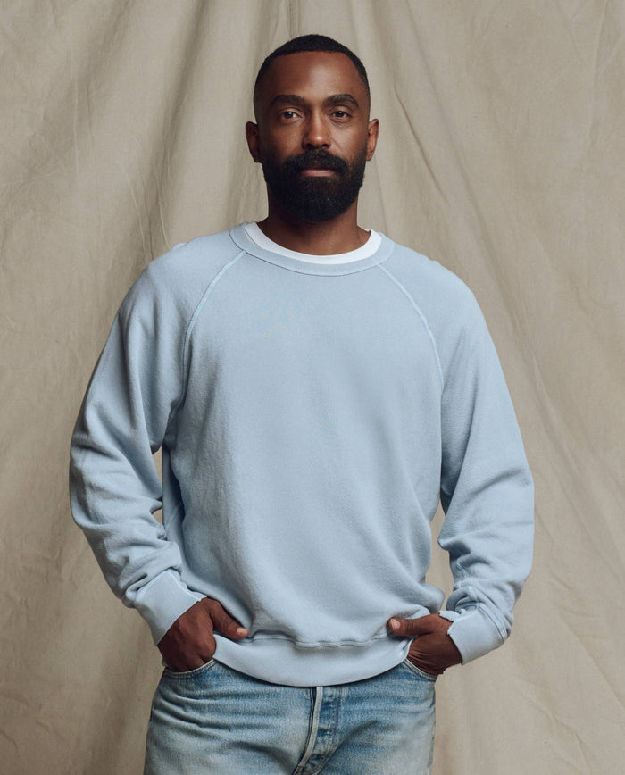 A bearded man stands before a beige fabric backdrop, wearing THE GREAT. SP26 SEASONAL CORE The Men's College Sweatshirt in Whisper Blue, a white undershirt, and light blue jeans with his hands in his pockets.