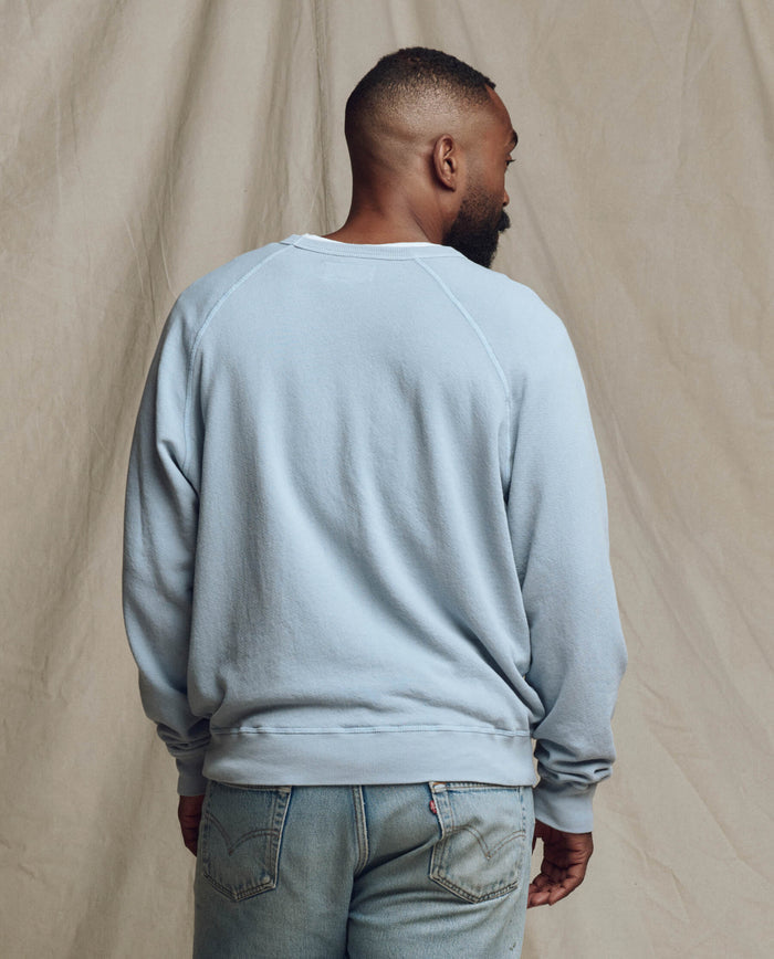 A man faces away, wearing THE GREAT.'s The Men's College Sweatshirt in Whisper Blue (SP26 SEASONAL CORE) and jeans, set against a neutral textured backdrop.