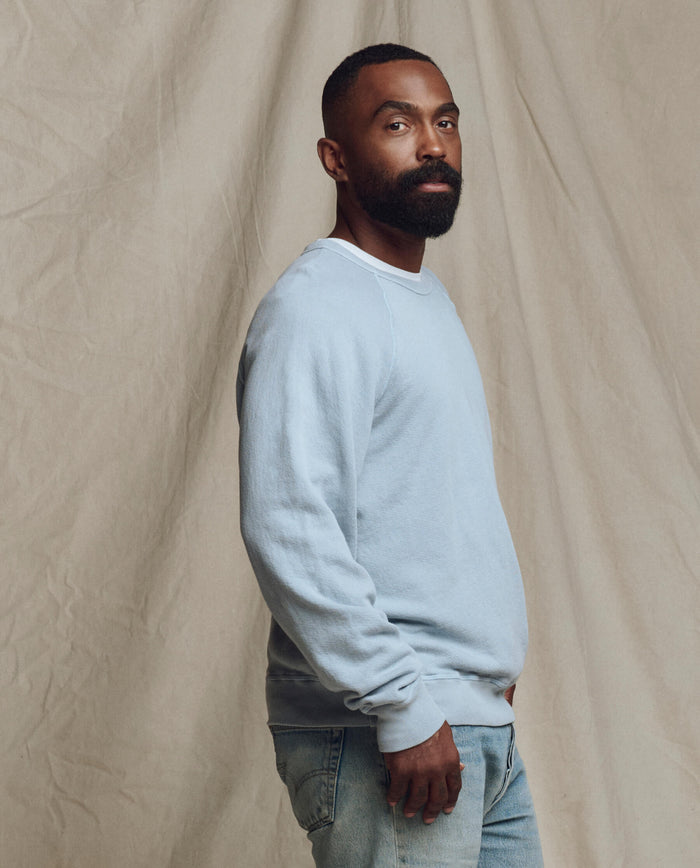 A bearded man stands sideways in front of a light beige backdrop, wearing THE GREAT. SP26 SEASONAL CORE The Men's College Sweatshirt in Whisper Blue and jeans.