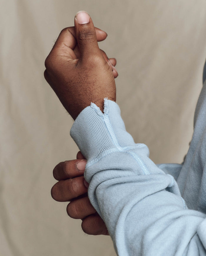 A person in THE GREAT. SP26 SEASONAL CORE The Men's College Sweatshirt -- Whisper Blue adjusts a cuff with a small tear at the seam.