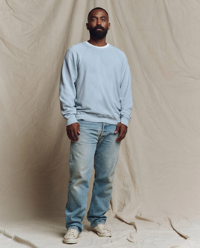 A bearded man stands against a beige fabric backdrop, wearing THE GREAT. SP26 SEASONAL CORE The Men's College Sweatshirt in Whisper Blue, paired with blue jeans and white sneakers.