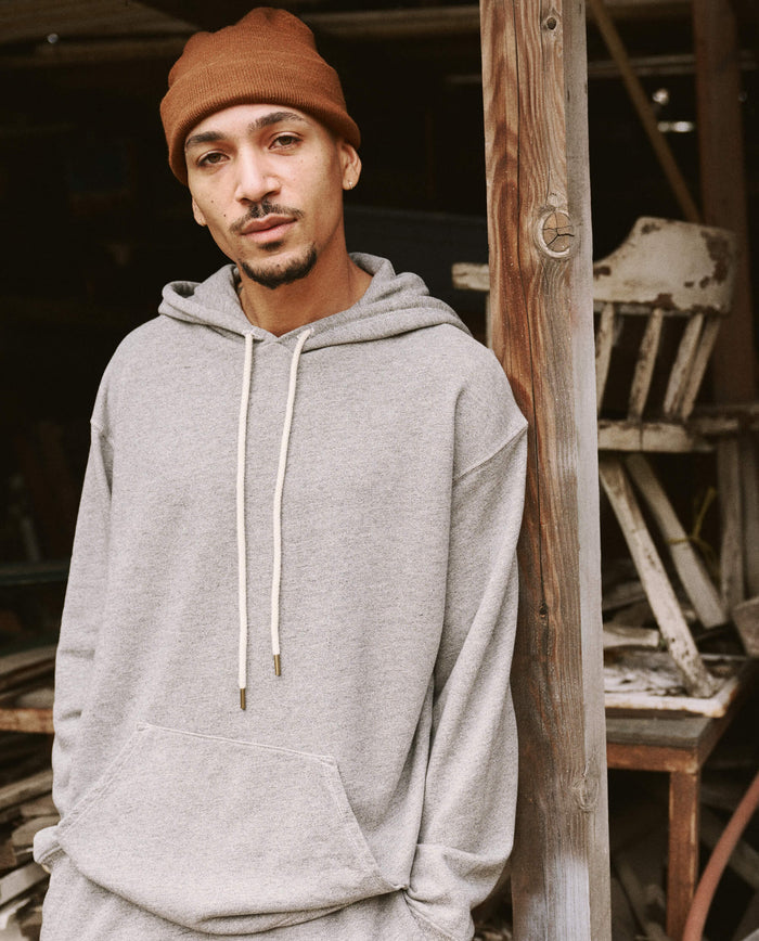 A man wearing The Men's Gym Drawstring Hoodie in Varsity Grey from THE GREAT. SU22 MENS and a brown beanie, both made in the U.S.A., stands next to a wooden post in an outdoor setting.