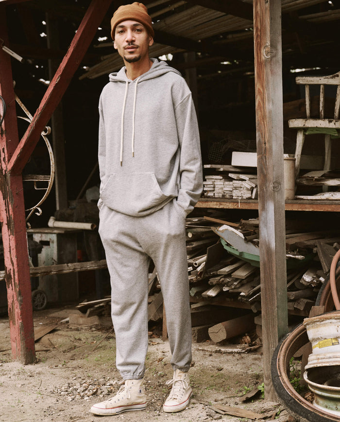 A man wearing THE GREAT. SU22 MENS' Varsity Grey Men's Gym Drawstring Hoodie, matching sweatpants, and white sneakers stands in a cluttered outdoor area with various wooden and metal objects around him.