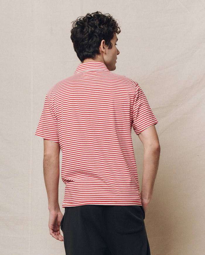 A person stands against a plain background wearing THE GREAT. SP25 POLO LOUNGE MEN's The Men's Lacrosse Polo in Sun Cherry Stripe, complemented by dark pants, facing away from the viewer.