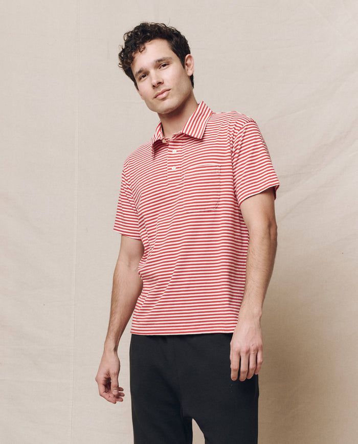 A person is wearing The Men's Lacrosse Polo in Sun Cherry Stripe with black pants, standing against a plain background.